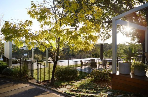 Sunlit landscaped outdoor area with a fenced swimming pool, paved patio, pergola-like white structural frame, raised timber planter or seating edge, mature trees, and garden planting. The setting appears residential and suburban, viewed from an adjacent paved outdoor area.