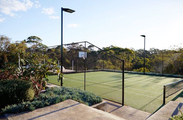 Bright outdoor view of a fenced hard-court sports area, likely a tennis or multipurpose court, with a central net, basketball hoop, tall black lighting poles, landscaped planting, and surrounding trees. The court has green synthetic surfacing and concrete walkways around the perimeter.