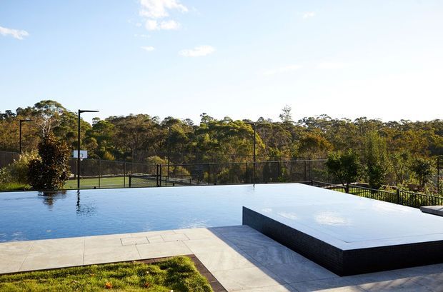 An outdoor pool area with a large rectangular swimming pool and raised dark-clad platform or spa, surrounded by pale paved terraces. Black fencing, slim outdoor lighting, landscaped lawn, and dense woodland beyond create a modern landscaped recreational setting under a bright sky.