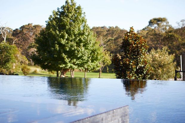 An outdoor infinity-edge swimming pool occupies the foreground, reflecting the surrounding trees and clear sky. Beyond the pool is a landscaped rural garden with mature deciduous and evergreen trees, lawn, and wooded hills in the background. The image has a calm, natural setting and no visible building.
