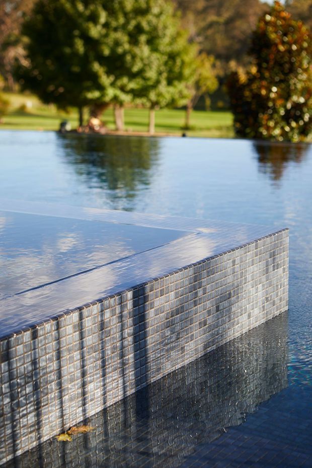 Close-up architectural photograph of a tiled infinity-edge swimming pool, showing a raised rectangular pool wall clad in small blue-grey mosaic tiles and water flowing over the edge. A landscaped garden with mature trees is softly blurred in the background.
