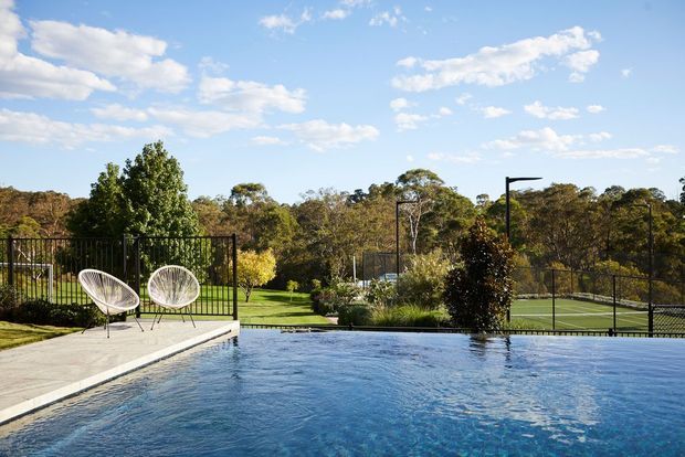 A landscaped outdoor pool area with a large blue swimming pool in the foreground, a pale paved deck, two woven lounge chairs, black metal fencing, lawn and mature trees beyond. A fenced tennis court is visible in the background under a bright blue sky.