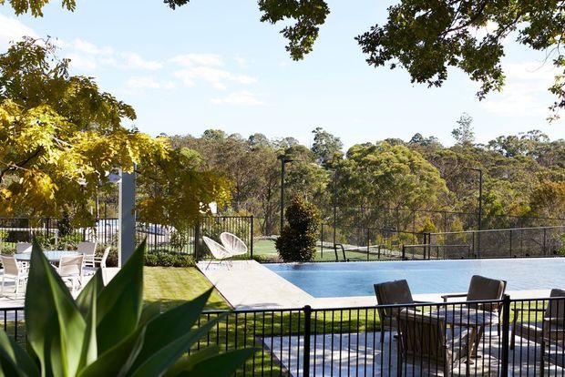 A landscaped outdoor pool area with a rectangular swimming pool, paved pool deck, outdoor dining furniture, black metal balustrades and fencing, and surrounding mature trees and planting. The setting appears leafy and suburban, with a bright open-air atmosphere.