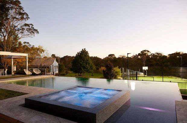 A landscaped outdoor leisure area with a large swimming pool and raised square spa in the foreground. A covered outdoor seating structure with hanging chairs sits beside the pool, while a fenced tennis or basketball court and mature trees extend into the background. The scene has a refined contemporary resort-like character in warm evening light.