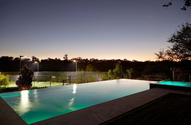 Twilight view of a large illuminated swimming pool with turquoise water, set beside a tiled deck and an adjacent fenced sports court. Trees and landscape form the background, with pool lighting creating a calm resort-like atmosphere.