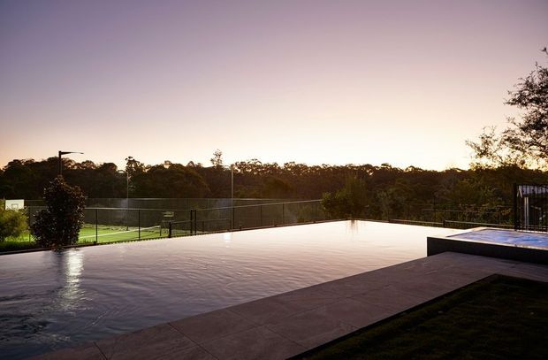 A modern outdoor pool terrace at dusk, featuring a broad reflective swimming pool, paved deck, raised spa or water feature at the right, dark metal fencing, and a wooded landscape beyond.