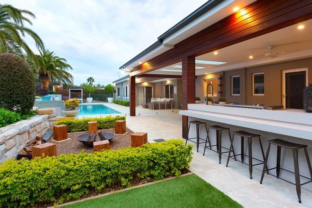 A luxurious contemporary outdoor entertaining area beside a swimming pool, featuring a long covered bar with integrated bench seating, timber-lined soffits, recessed lighting, and dark-framed openings. The space opens onto a landscaped garden with clipped hedges, palms, stone retaining walls, a fire-pit seating area, and a pool. A bright interior living or dining area is visible beyond the covered opening.