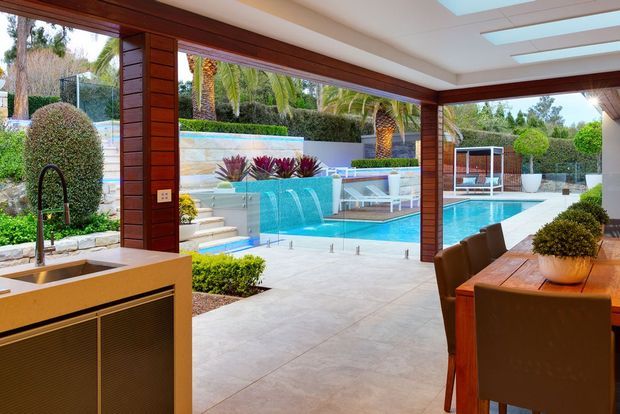 A refined open-plan indoor-outdoor entertaining space with a timber-framed glazed enclosure overlooking a landscaped swimming pool courtyard. The interior includes a compact kitchen or bar with sink, a long timber dining table, upholstered dining chairs, skylights, and warm timber wall framing. Outside are a turquoise swimming pool, water feature, paving, clipped hedges, and sculptural planting.