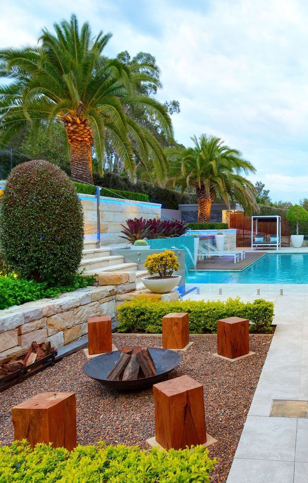 A landscaped tropical pool courtyard with a bright turquoise swimming pool, raised spa or water feature, stone retaining walls, clipped hedges, palms, ornamental planting, and a gravel outdoor entertaining area. A circular fire pit is surrounded by chunky timber stump seats, while pale paving leads around the pool.