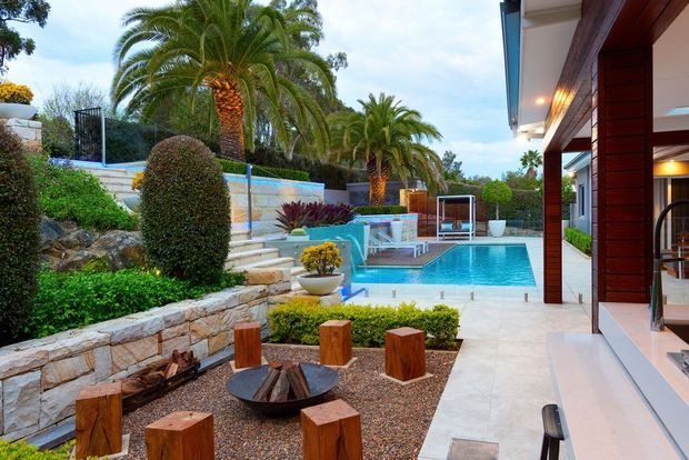 A landscaped luxury residential outdoor area viewed from a covered terrace, featuring a bright swimming pool, raised spa or water feature, stone retaining walls, manicured hedges, palm trees, and a gravel fire-pit seating area with timber stools. The adjacent house has warm timber-clad columns and large glazed openings.