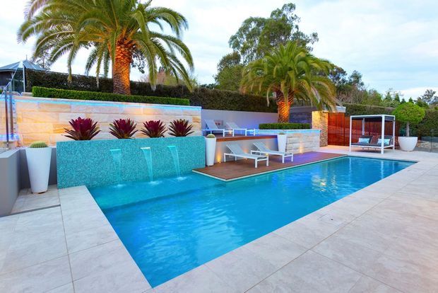 A landscaped outdoor pool area with a large rectangular swimming pool, integrated turquoise water feature wall, raised spa or planter elements, palms, tiled paving, built-in outdoor seating, and a covered poolside lounge. The design has a contemporary resort-like character with warm timber accents and illuminated water features.