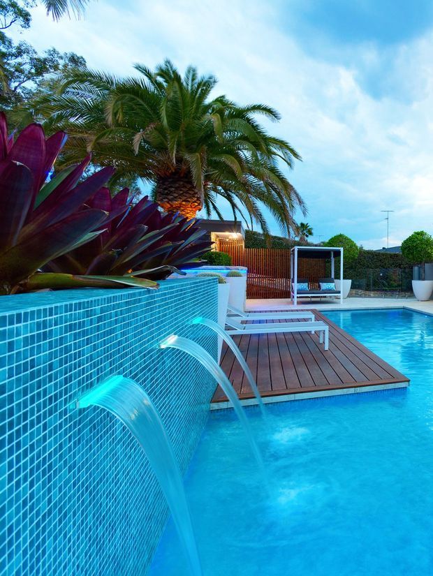 A tropical outdoor swimming pool with bright blue mosaic-tiled water features cascading from a raised wall into the pool. A timber deck with white loungers extends over the water, surrounded by palm trees, lush planting, privacy fencing, and a shaded cabana. The scene has a resort-like, vibrant outdoor setting.
