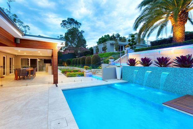 Contemporary residential outdoor entertaining area with a large bright-blue swimming pool, tiled water-feature wall with cascading spillways, landscaped garden beds, palm trees, and an open-plan dining pavilion with timber-lined soffits and glazed doors.