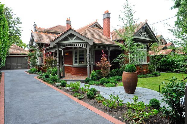 A landscaped front garden and driveway leading to an ornate red-brick period home. The house features multiple steep tiled gables, decorative timber bargeboards and fretwork, covered verandahs, brick chimneys, and a detached-looking garage integrated at the left. Mature planting, clipped lawn, paved paths, and a large terracotta planter complete the exterior scene.