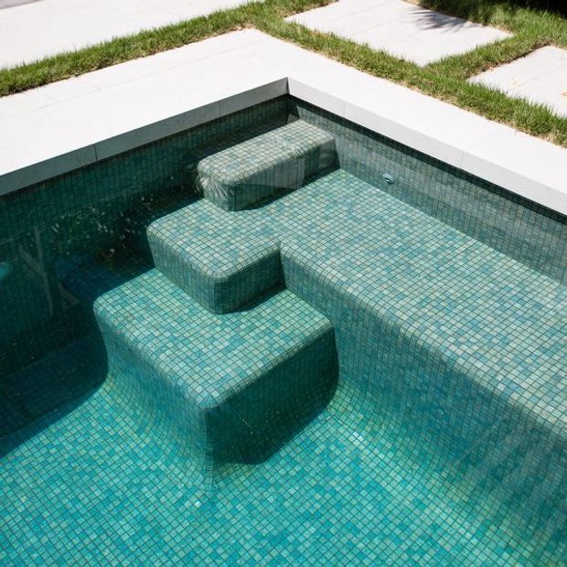 Close-up view of a contemporary swimming pool with integrated submerged bench seating and stepped platforms, fully finished in small turquoise and teal mosaic tiles. Pale concrete coping and rectangular concrete pavers surround the pool, with narrow strips of grass between them.