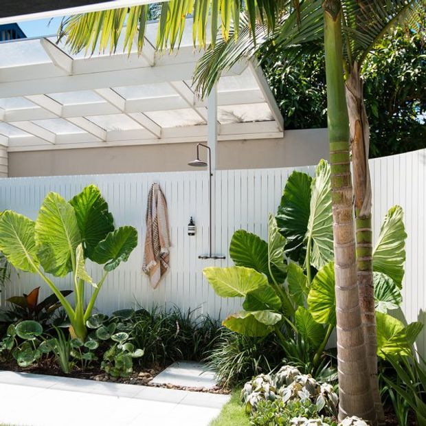 Bright tropical outdoor shower area beside a white vertical-panel fence, with a palm tree, lush broad-leaf planting, a paved stepping path, and a translucent roof/pergola structure overhead. The space has a fresh, relaxed resort-like atmosphere.