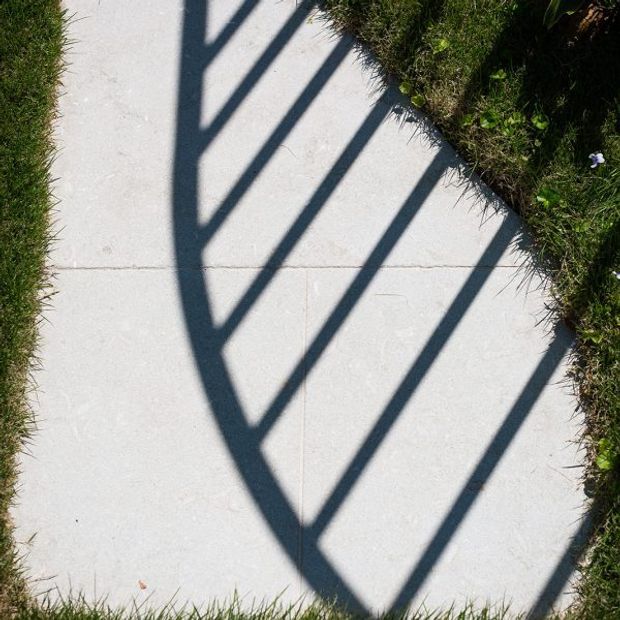 A close-up, top-down view of a pale concrete or stone-paved outdoor walkway bordered by grass. Strong dark linear shadows from a railing or balustrade create a repeating geometric pattern across the paving.