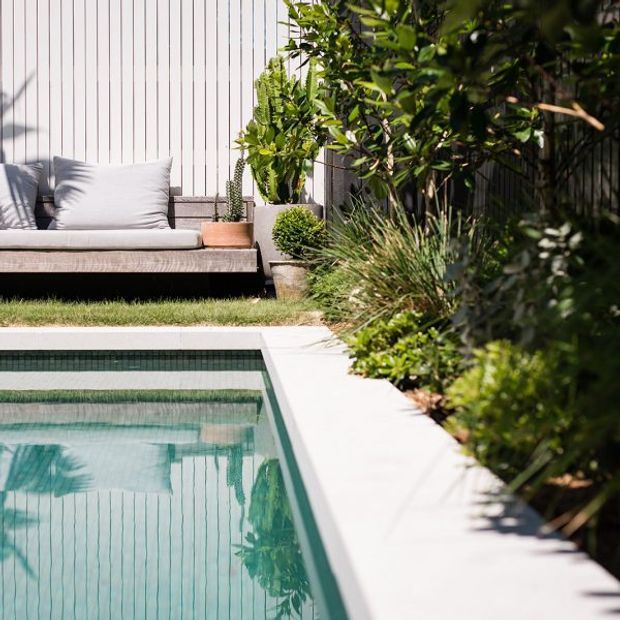A landscaped residential pool area with a rectangular swimming pool edged in pale paving. Dense planting borders one side, while a built-in outdoor lounge with light cushions sits against a vertical timber-lined privacy wall. The scene has a clean, contemporary garden design with strong reflections in the water.