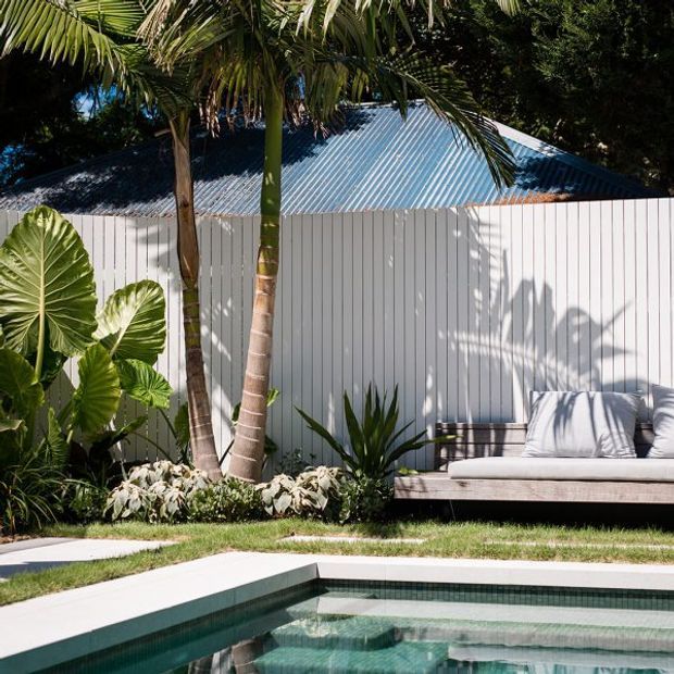 A tropical landscaped pool area beside a white vertical-panel building with a corrugated metal pitched roof. Palm trees and lush foliage frame a built-in outdoor lounge bench with cushions, while the foreground shows the edge of a rectangular swimming pool and pale paving.