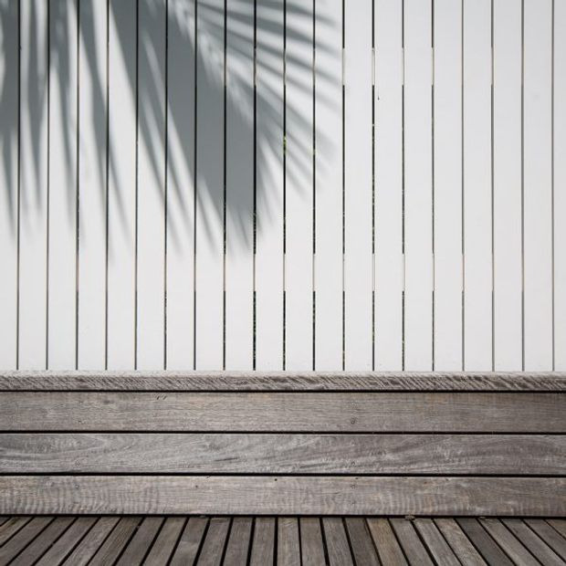 Close-up architectural detail of a minimalist outdoor deck or balcony, featuring weathered horizontal timber-clad raised seating or planter-like structure, dark timber decking, and a white vertical slatted wall casting strong linear shadows.