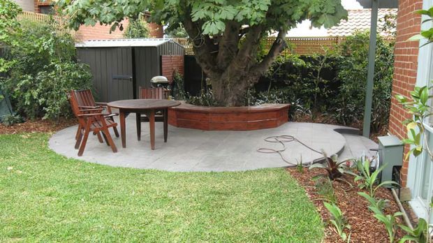 A landscaped residential courtyard garden with a curved paved patio, outdoor timber dining table and chairs, a raised curved brick garden bed beneath a mature tree, and a dark garden shed. Brick walls, dense planting, lawn, and a simple outdoor seating area create a private backyard setting.