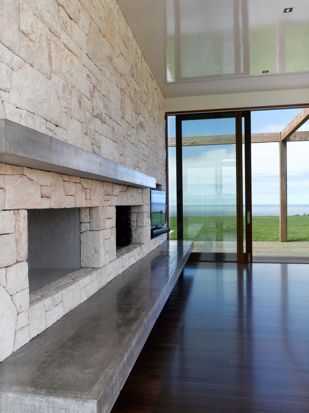 A refined living room centered on a large rough-cut stone fireplace with a broad concrete hearth and mantel. Dark timber flooring, extensive floor-to-ceiling glazing, and slim dark window and door frames open toward a grassy coastal landscape and ocean beyond. The room has a high ceiling with recessed downlights and a bright, minimal architectural character.
