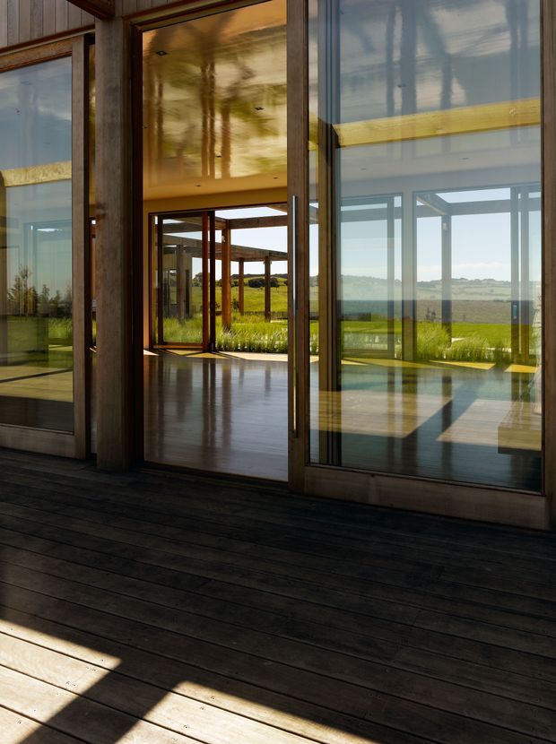 A modern architectural interior or covered outdoor threshold with dark timber decking, substantial timber-framed glazing and a glazed doorway. The highly reflective glass opens views toward a sunlit rural landscape, while warm timber ceiling and structural posts create a strong indoor-outdoor connection.