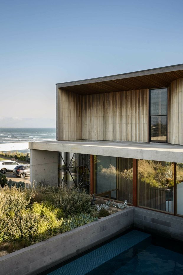 A modern multi-level coastal house with a flat-roofed, cantilevered concrete structure, vertical timber cladding and a deep timber-lined balcony or terrace. Floor-to-ceiling glazing overlooks a narrow swimming pool, native-style landscaping and a visible beach and ocean beyond. The composition highlights the building exterior and its relationship to the coastal site.