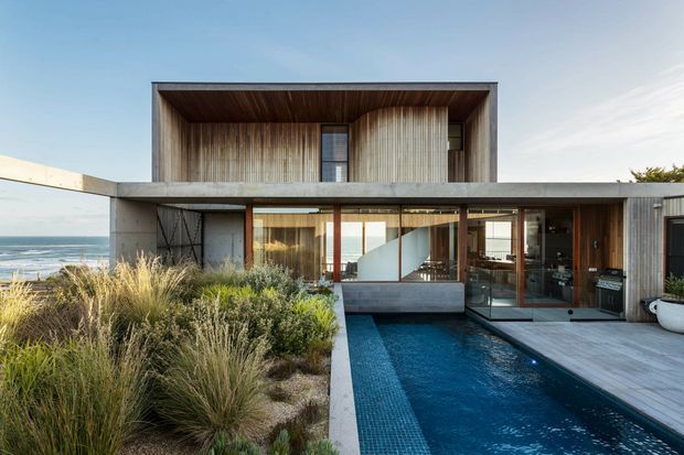 A contemporary two-storey coastal residence with a strong rectilinear composition, deep recessed upper-level terrace or balcony, extensive glazing, timber-lined soffits, and concrete surfaces. The home overlooks the ocean and is surrounded by drought-tolerant grasses and coastal planting. A narrow swimming pool runs alongside a paved outdoor area and glazed living spaces.