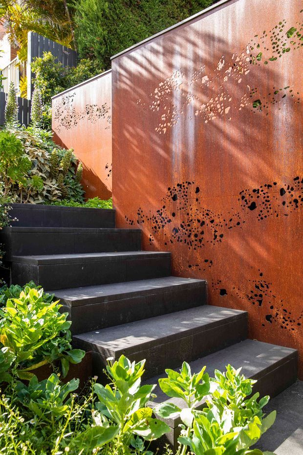 A landscaped outdoor stairway rises between dark tiled or stone steps and a tall rusted corten-steel screen wall with decorative perforated patterns. Dense green planting softens the composition, while sunlight creates leafy shadows across the warm metal surface.
