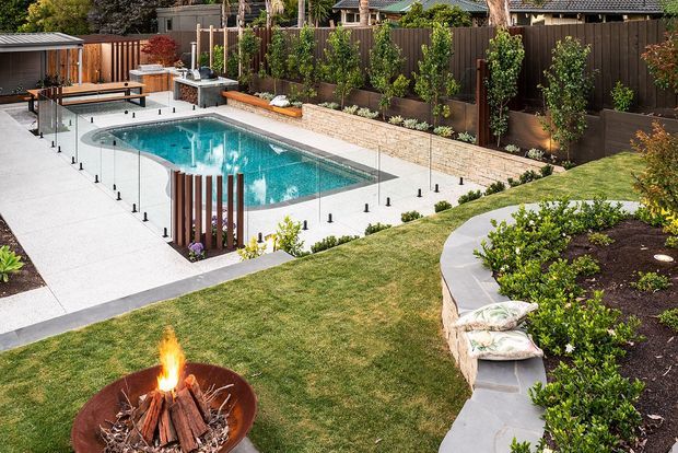 Landscaped backyard with a turquoise swimming pool enclosed by frameless glass fencing, surrounded by pale concrete paving, timber screening, raised garden beds, lawn, and a fire pit in the foreground.