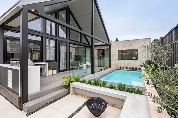 Contemporary courtyard-style home exterior with a dramatic gabled glazed pavilion opening onto a timber deck and compact swimming pool. The dark-framed floor-to-ceiling glazing, charcoal structural framing, light brick walls, glass balustrade, built-in concrete garden edging and outdoor fire bowl create a refined indoor-outdoor entertaining space.