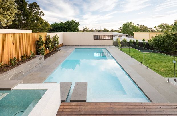 A contemporary landscaped backyard centered on a long rectangular swimming pool, with a smaller spa in the foreground, timber decking, pale paved surrounds, rendered boundary walls, timber screening, planting, and a glass pool fence. The scene is bright and open with a clean, refined outdoor design.