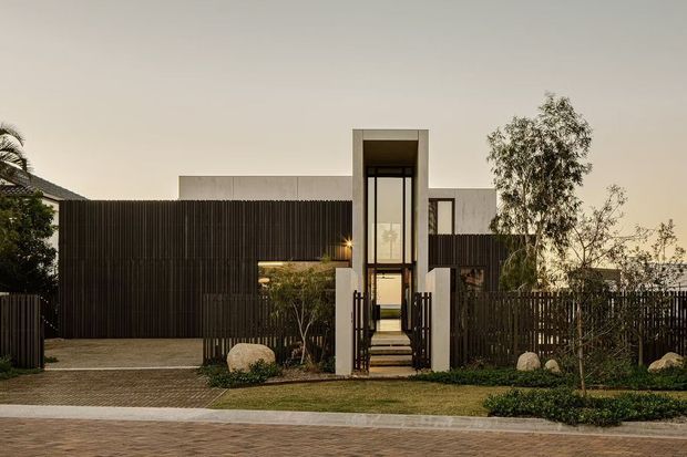 Contemporary residential facade viewed from the street at dusk, featuring dark vertical screening, a central glazed entry tower with a tall rectangular portal, pale rendered or concrete elements, landscaped planting, and a paved driveway and footpath.