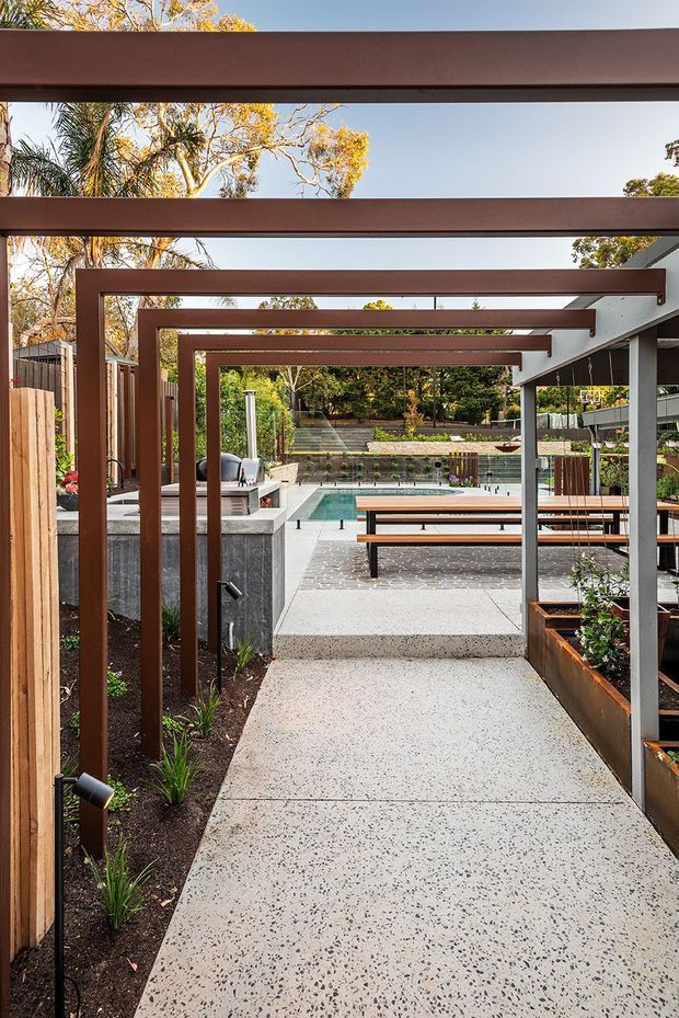 A landscaped outdoor courtyard and pool area viewed along a speckled concrete walkway beneath a long sequence of dark timber or metal pergola beams. The space includes raised planter beds, built-in benches and tables, a pool, stepped garden terraces, timber fencing, and a contemporary pavilion-like structure.