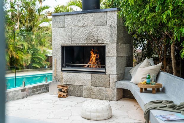 Outdoor entertaining area featuring a substantial stone-clad fireplace with a live fire, built-in concrete or rendered bench seating, a small timber side table, pale irregular-paved flooring, and a swimming pool visible beyond the glass balustrade. Lush tropical landscaping surrounds the space.