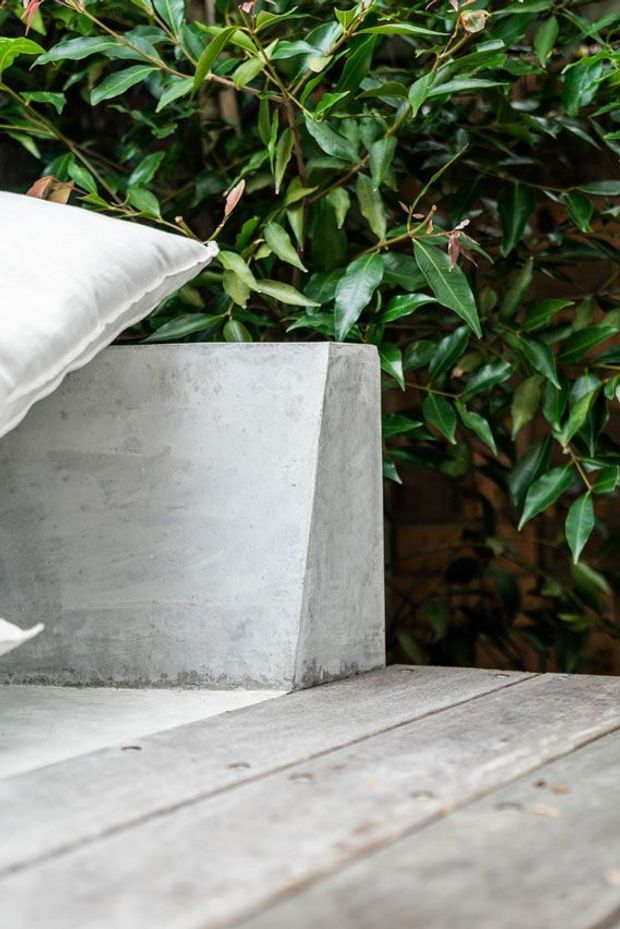 Close-up outdoor scene featuring a weathered timber deck or tabletop, a raw light-grey concrete planter or bench, and a white cushion, set against dense glossy green foliage. The image has a natural, tactile, contemporary garden atmosphere.