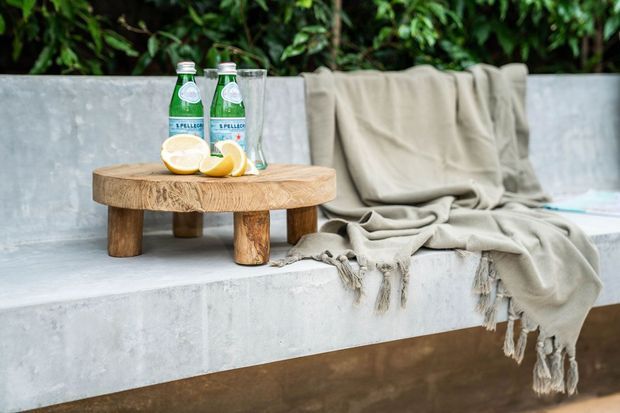 A styled outdoor concrete bench or ledge with a draped beige fringed throw, a low round natural timber side table, sparkling water bottles, glasses, and lemons. Dense greenery forms the background, creating a relaxed garden or patio setting.