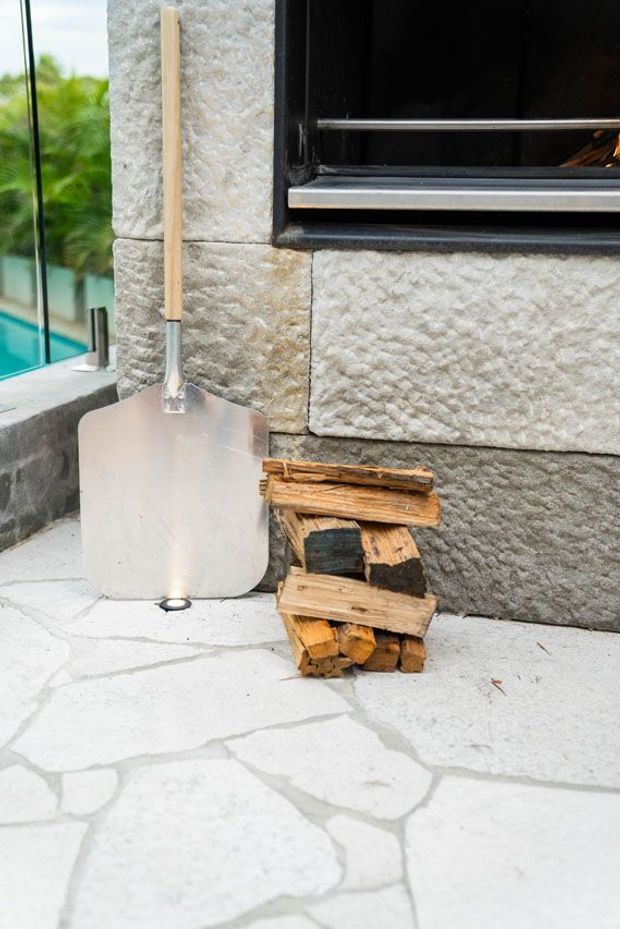 Close-up of an outdoor area beside a modern building, showing a metal shovel with a wooden handle leaning against a textured stone-clad wall, a small stack of firewood, pale irregular stone paving, and part of a dark-framed window. A pool or turquoise outdoor surface is visible at the left edge.
