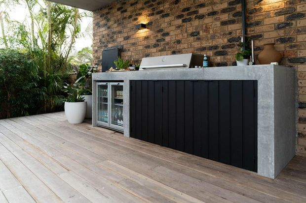 An outdoor entertaining area with a built-in barbecue kitchen and drinks refrigerator. The island has dark vertical-panel cabinetry, a grey concrete benchtop and side panels, and sits against an exposed brick wall. Timber decking, lush screening plants, integrated lighting, and a sheltered roof create a warm, practical alfresco setting.