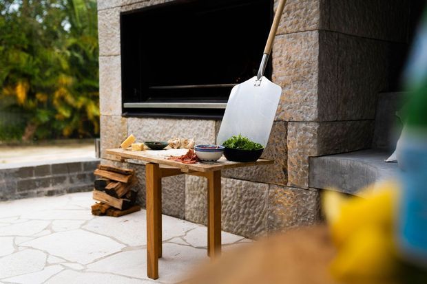 Outdoor cooking area with a built-in masonry fireplace or grill clad in textured stone, a wooden preparation table holding bowls and ingredients, stacked firewood, and pale irregular stone paving. Lush vegetation is visible in the background, creating a rustic, natural setting.