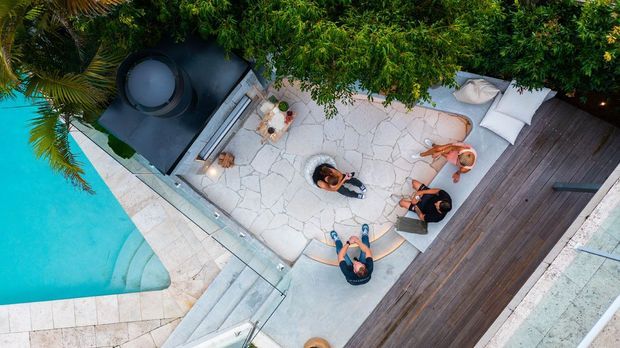 Overhead view of a landscaped outdoor entertainment area beside a turquoise swimming pool. The space includes pale irregular stone paving, a dark timber deck, a large black outdoor fireplace or built-in feature, glass pool fencing, and people gathered around outdoor seating.