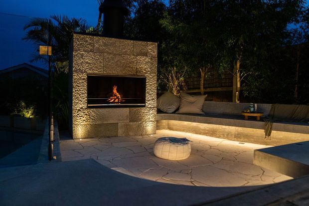 A moody outdoor entertaining area at dusk, centred on a built-in textured masonry fireplace with a lit fire. A long cushioned bench wraps along the rear boundary, with warm under-seat lighting, a low round ottoman, paved flooring, and surrounding landscaping and trees.
