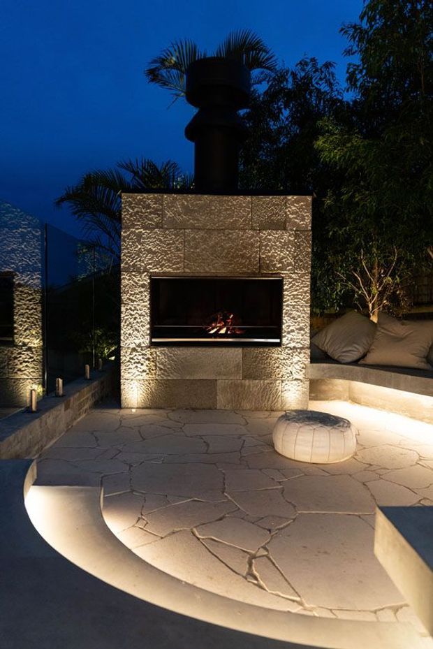 A moody outdoor entertaining area at dusk featuring a built-in rectangular fireplace clad in textured stone, with a dark metal chimney. The fireplace is set within a curved stepped patio with integrated warm lighting, surrounded by lush planting and built-in bench seating with cushions. A small rounded upholstered ottoman sits on the patterned stone paving.