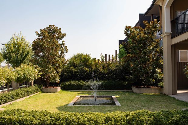 A landscaped courtyard garden beside a substantial multi-storey residential building, featuring clipped hedges, mature trees and shrubs, lawn, raised planting beds, and a small rectangular central fountain. The building has cream rendered walls, dark balcony railings, and a covered architectural colonnade at right.