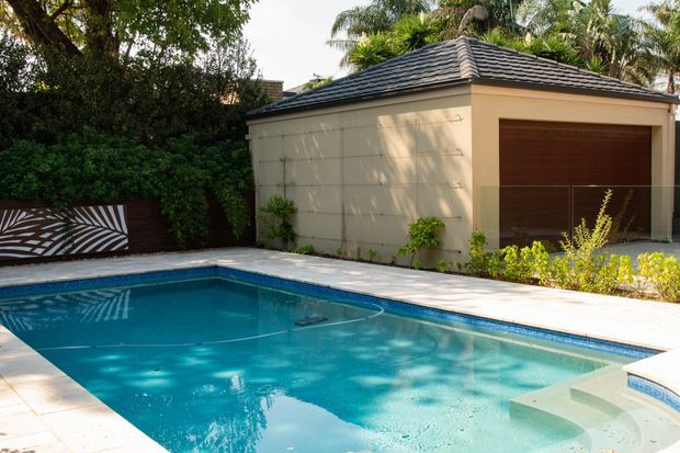 A landscaped residential pool area beside a detached garage or pool house. The rectangular swimming pool has pale paving, integrated steps, and a shallow lounging area. Behind it is a cream rendered structure with a dark tiled hipped roof and a large brown timber-look garage door, bordered by lush tropical planting and a decorative patterned privacy gate.