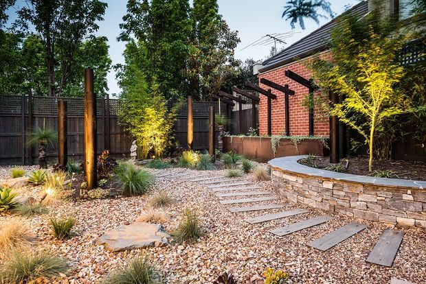 A landscaped residential courtyard or side garden with a gravel ground plane, stepping-stone path, layered stone retaining wall, native grasses, small ornamental trees, timber posts and dark screening fences. A red-brick building with a pitched dark roof forms the backdrop.