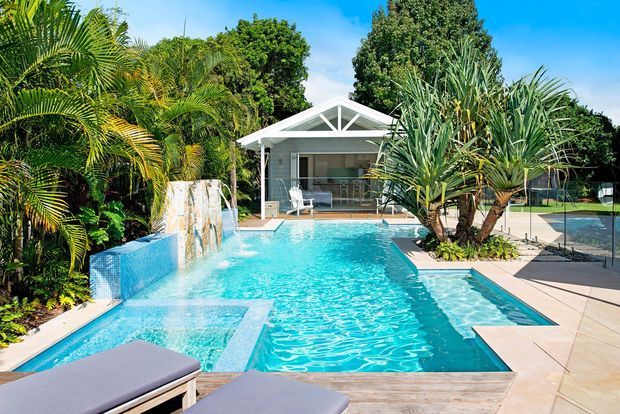 Bright tropical outdoor pool area with a large turquoise swimming pool, integrated shallow ledges and water features, surrounded by lush palms and planting. A small contemporary pavilion with a glazed gable-front facade sits beyond the pool, while paved terraces, loungers and a fenced recreational court occupy the surrounding space.