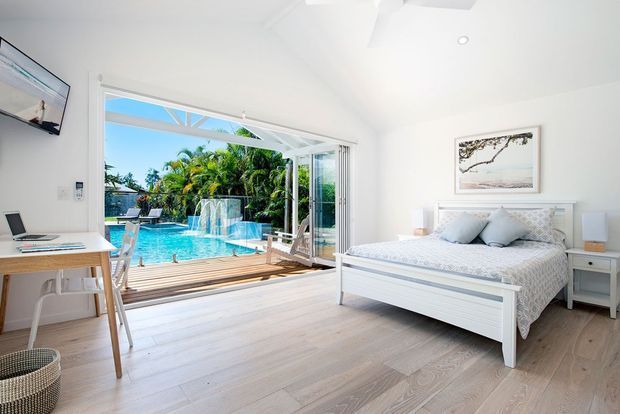 Bright coastal-style bedroom with a white panelled bed, pale timber flooring, white walls, and a compact desk. Large open bi-fold glazing connects directly to a timber deck and a turquoise swimming pool, with lush tropical planting beyond. The room is airy and uncluttered, with a ceiling fan, bedside tables, and soft blue-grey bedding.