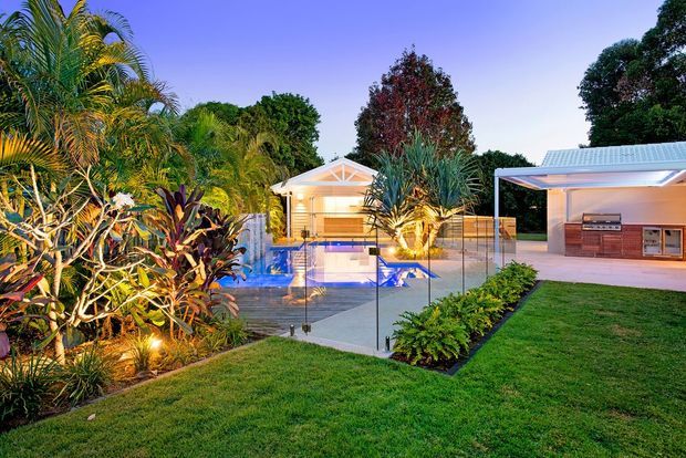 A landscaped residential backyard at dusk featuring a brightly lit swimming pool enclosed by glass fencing, tropical planting, a small pool pavilion, and a covered outdoor entertaining area with a built-in barbecue and cabinetry. The scene has a relaxed resort-like character with illuminated gardens and lawn.